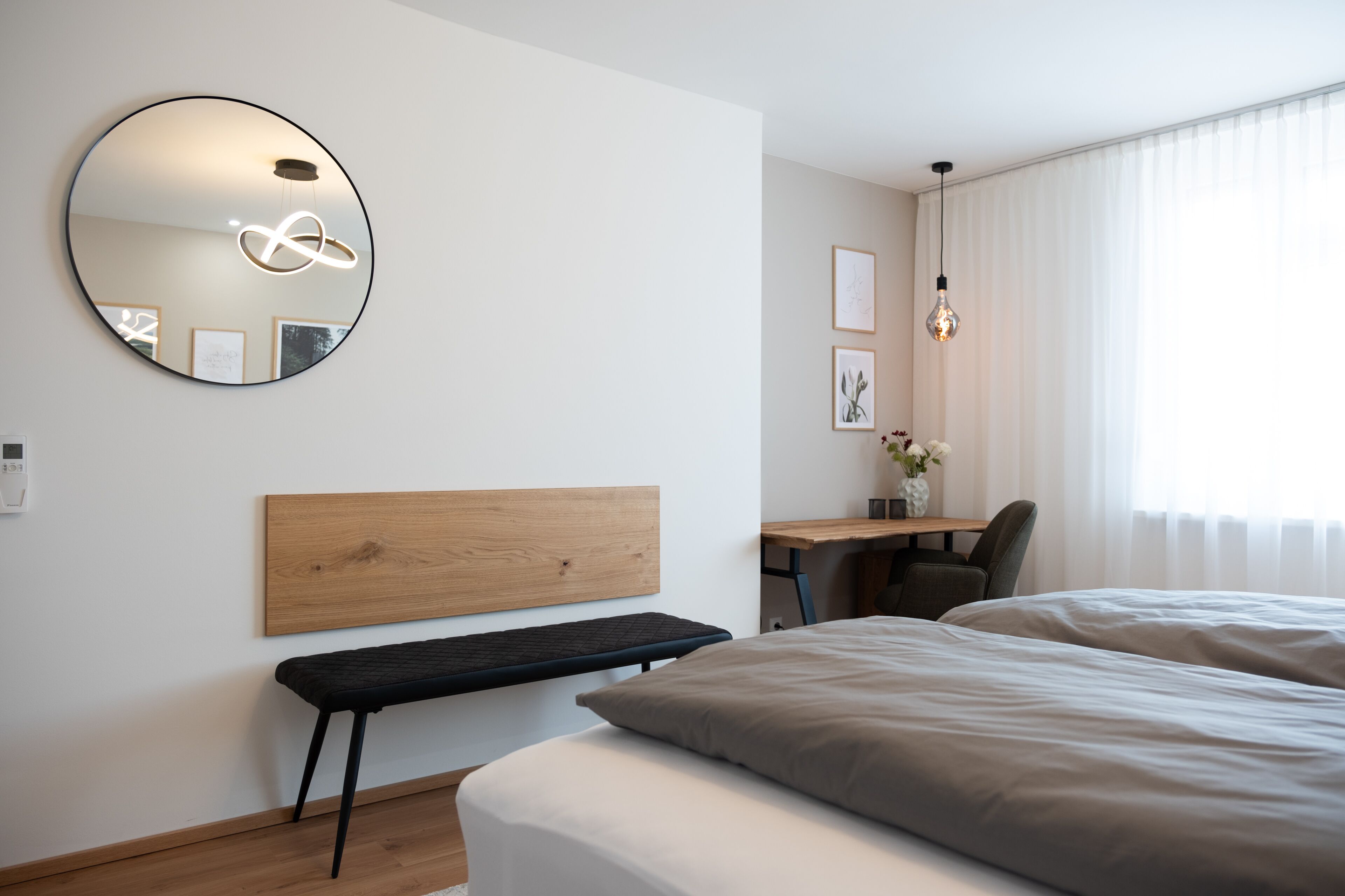 A modern hotel room with a wooden headboard, a round mirror, a desk with a chair, and a large bed.