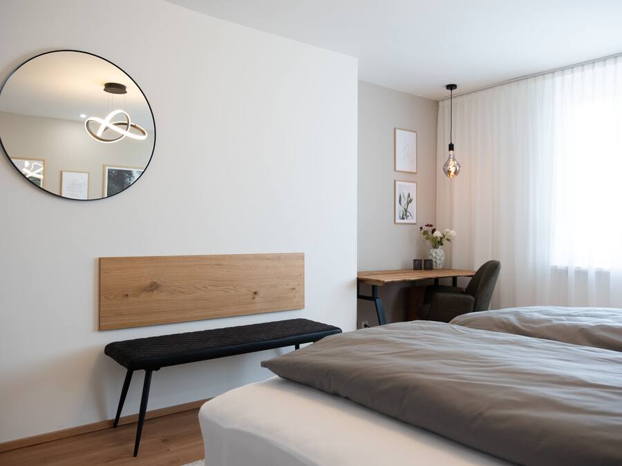 A modern hotel room with a wooden headboard, a round mirror, a desk with a chair, and a large bed.