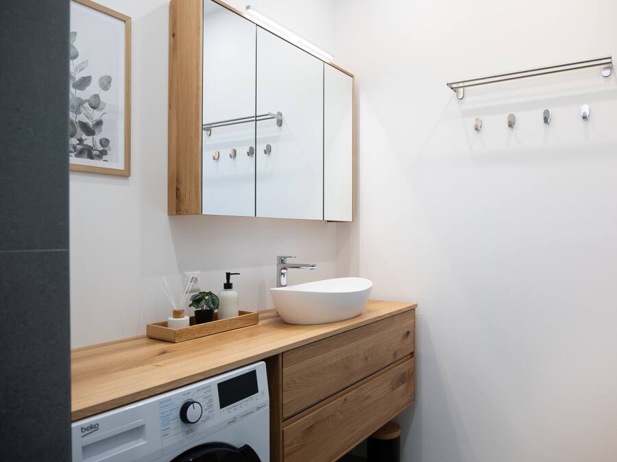 A modern bathroom with a wooden vanity, a white sink, a mirror, and a washing machine.