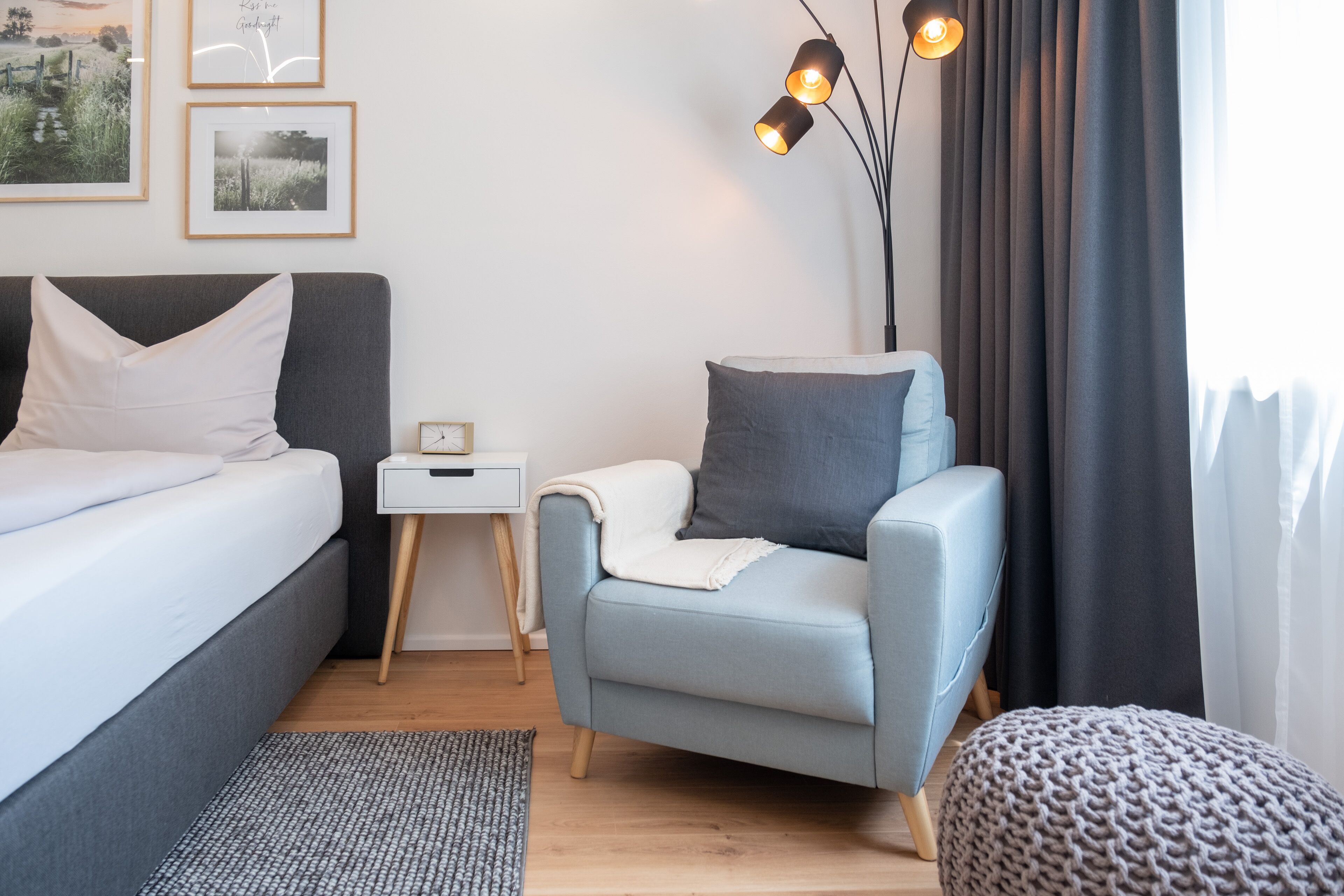 Et moderne soveværelse med en seng, et lille natbord, en lyseblå lænestol, en strikket skammel og et sengebord med lampe.