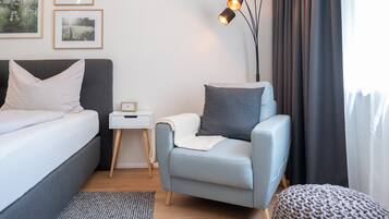 Et moderne soveværelse med en seng, et lille natbord, en lyseblå lænestol, en strikket skammel og et sengebord med lampe.