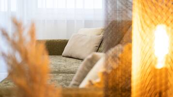 Un salon chaleureux avec un canapé et des coussins, une fenêtre avec des stores et une plante décorative au premier plan.