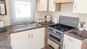 A modern kitchen with a gas stove, oven, and sink. There are wooden cabinets, a window with blinds, and a welcome sign on the countertop. - 3 Bed New Lodge, Sleeps 4, Pets Welcome (Scunthorpe)
