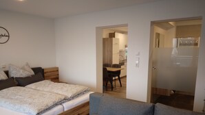 Un dormitorio moderno con cama de madera, sofá gris y un pequeño comedor visible a través de una puerta abierta.
