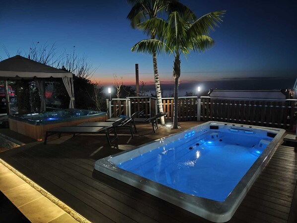 A hot tub and lounge chairs on a wooden deck with a view of the ocean at sunset.
