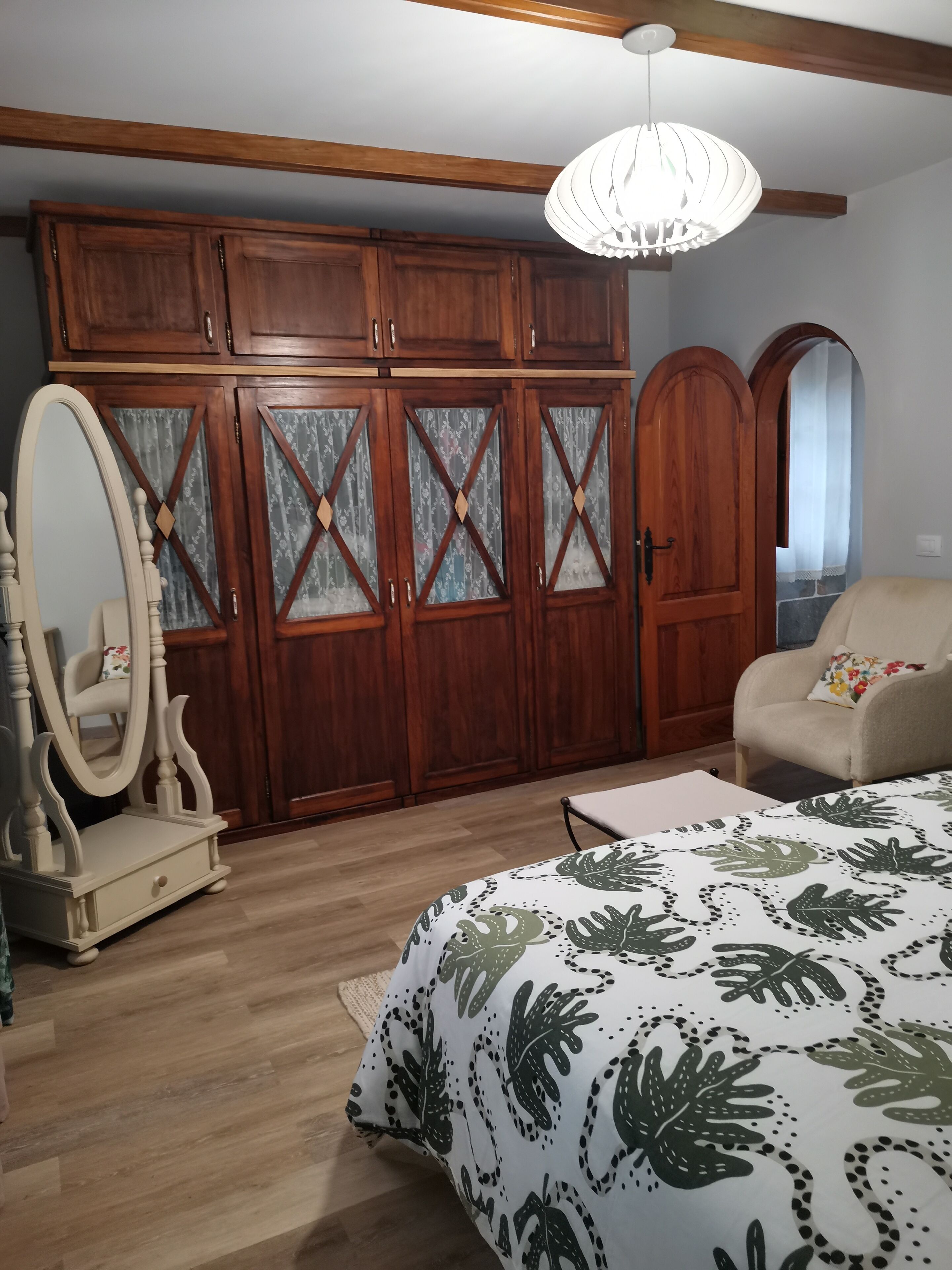 A bedroom with a wooden wardrobe, a mirror, a bed with a leaf-patterned duvet, and a hanging light fixture.
