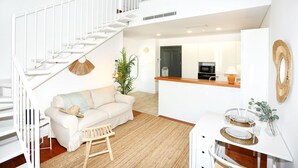 Un intérieur moderne avec un escalier blanc, un espace de vie comprenant un canapé et une table basse, une table à manger dressée pour deux personnes, et une cuisine en arrière-plan.