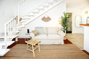 A modern living room with a white staircase, a white sofa with cushions, a small wooden stool, a woven rug, and a potted plant. - Watermelon Sugar Apartment (Faro)