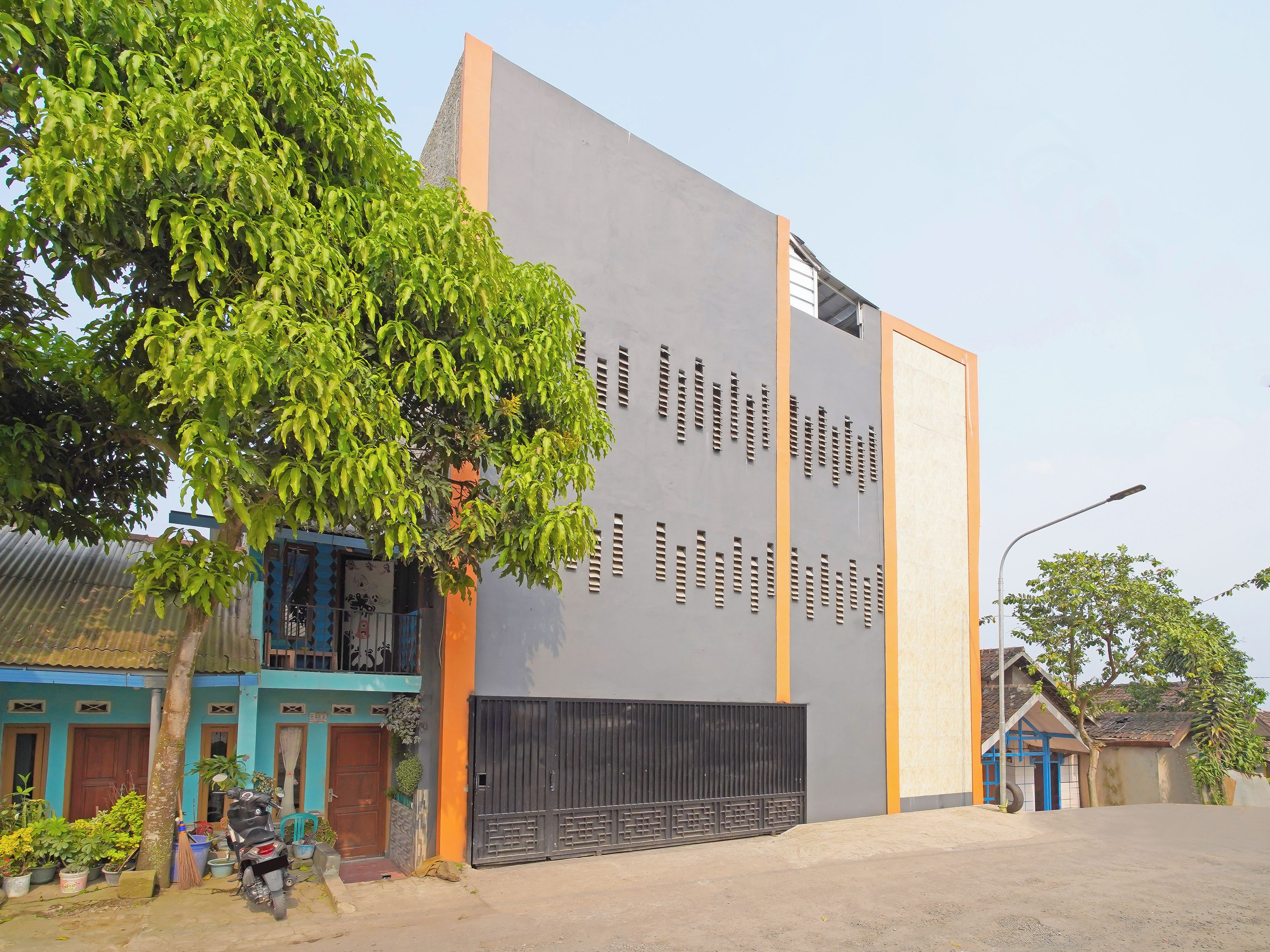 A modern building with a grey facade and orange accents, featuring a large black gate and a tree in the foreground.