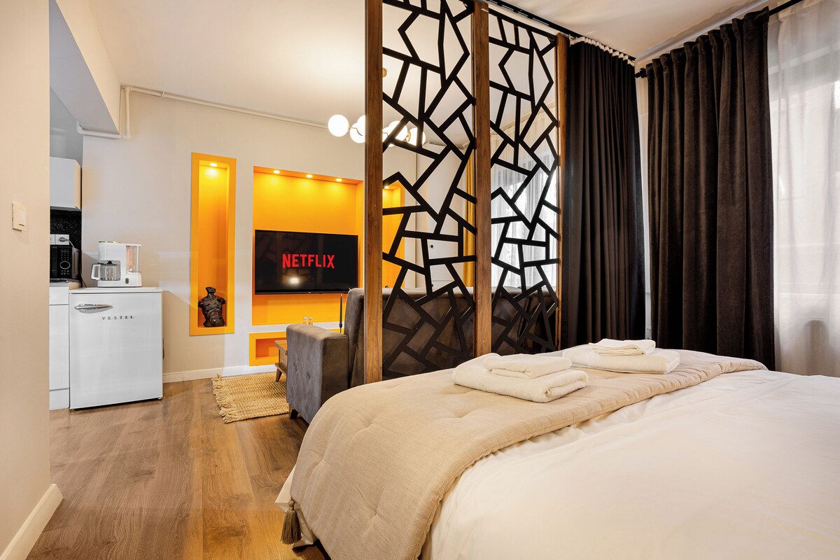 A modern hotel room with a large bed, a desk with a coffee machine, a TV, and a decorative partition.