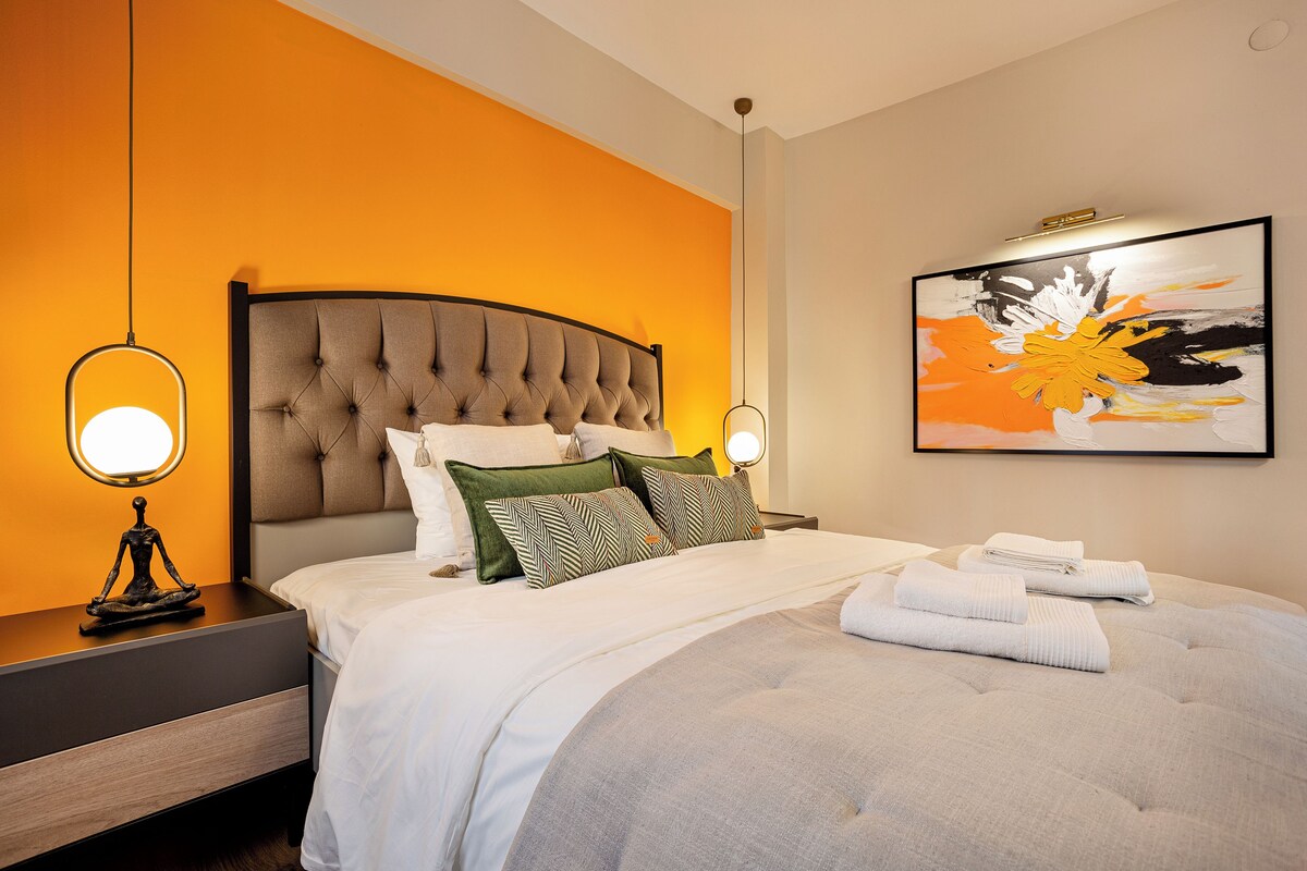 A modern bedroom with a tufted headboard, a large bed with white bedding, two bedside lamps, and a framed abstract painting on the wall.