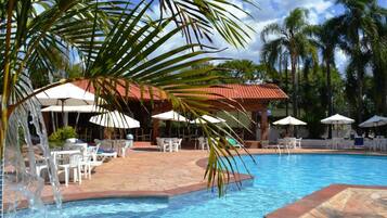 A swimming pool with lounge chairs and umbrellas, surrounded by palm trees and a red-roofed building.