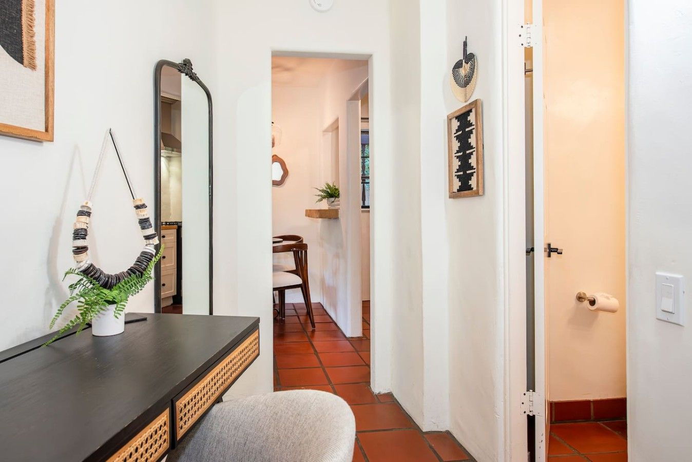 A hallway with a mirror, a decorative plant, and a door leading to a room with a chair and a small table.