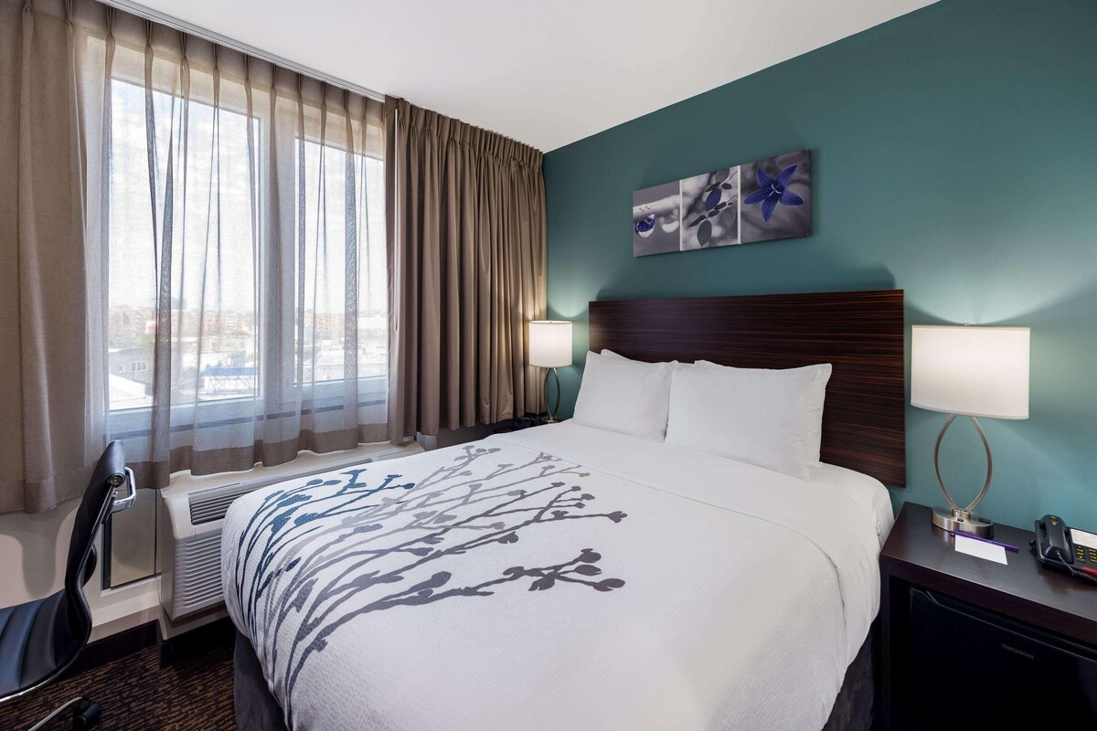 A hotel room with a bed, two bedside lamps, a window with curtains, a wall-mounted artwork, and a desk with a phone and a notepad.