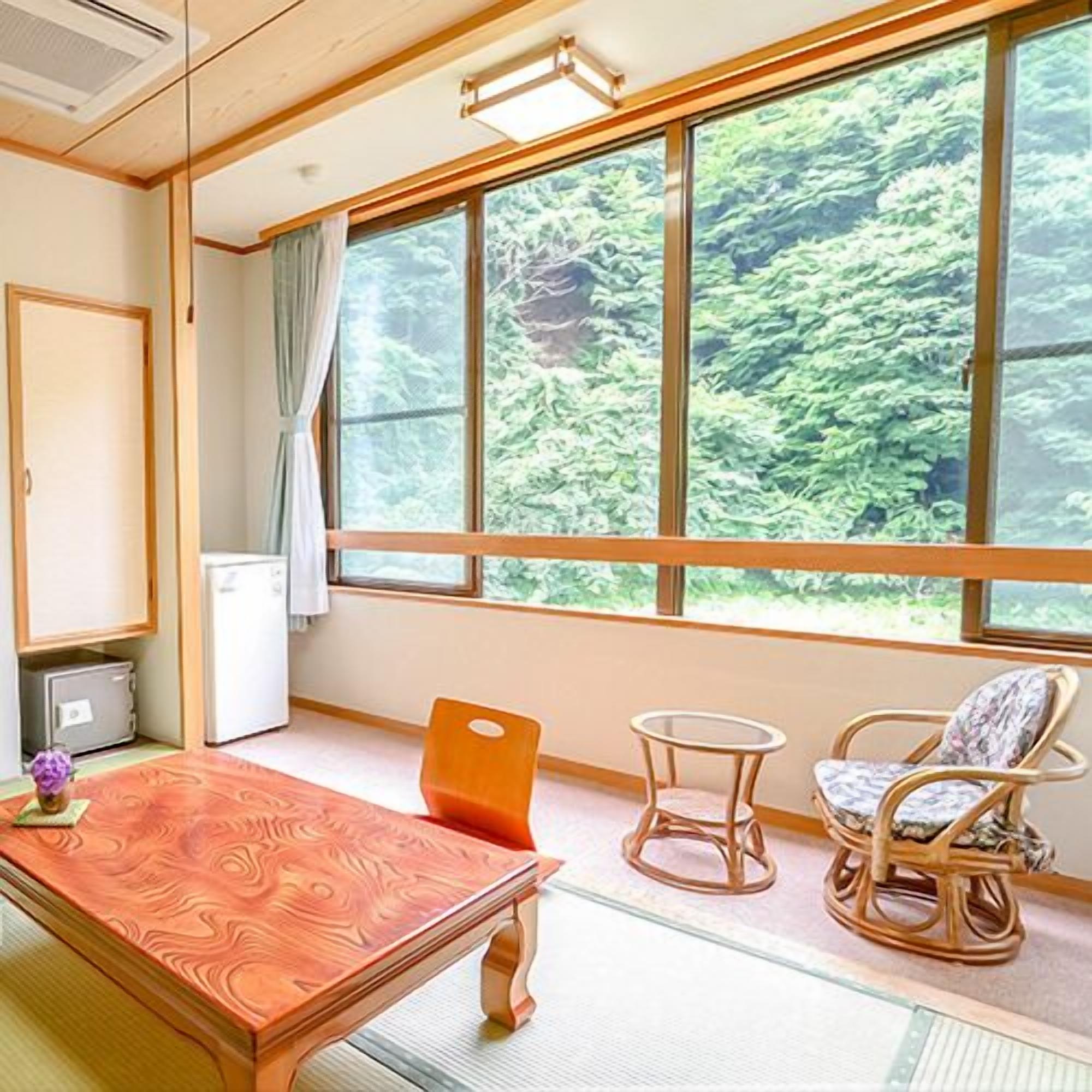 A room with a wooden table, two chairs, a small round table, and a view of greenery through large windows.