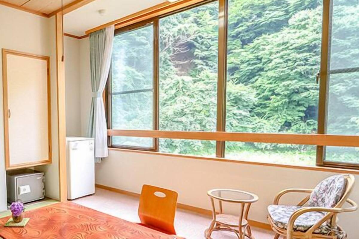 A room with a wooden table, two chairs, a small round table, and a view of greenery through large windows.