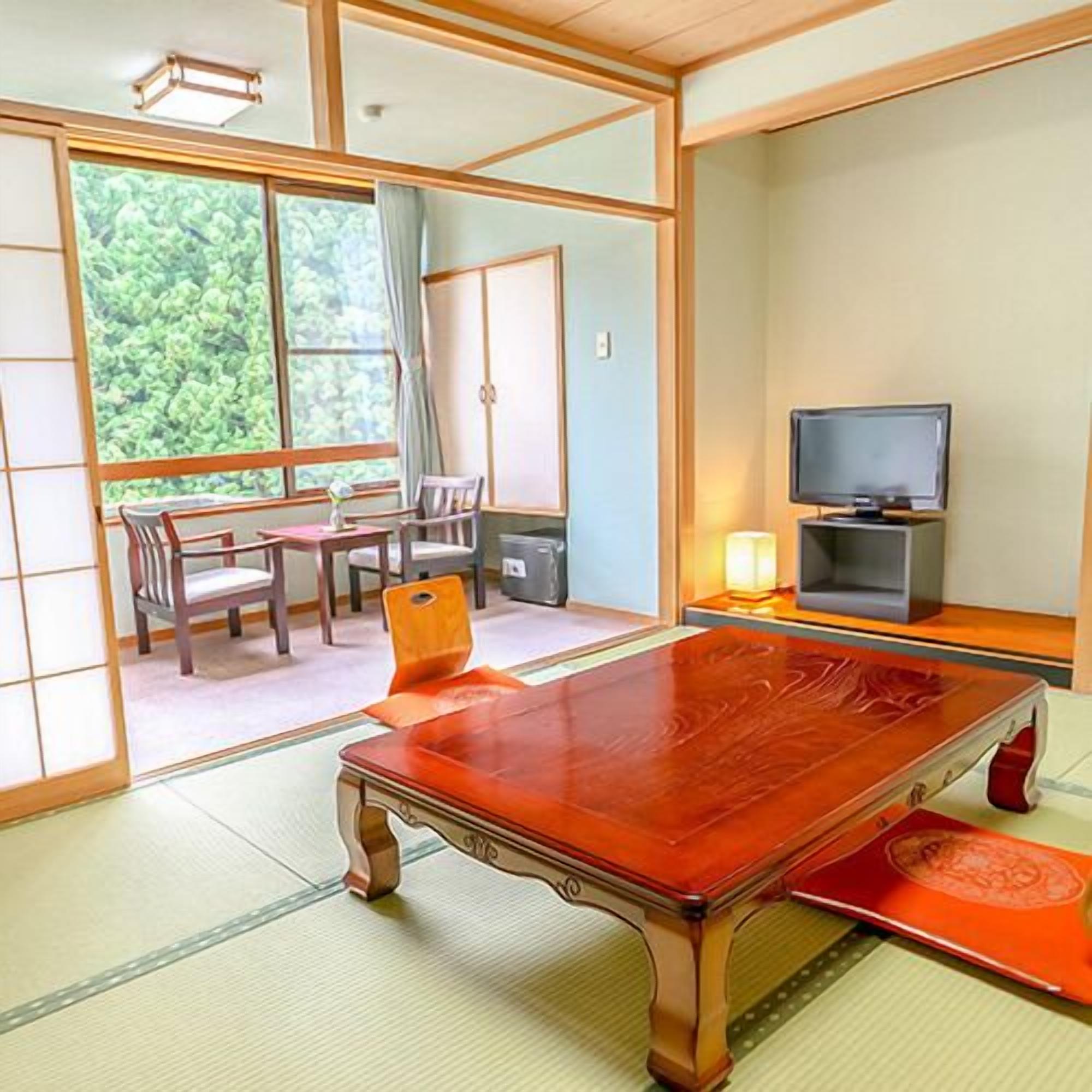 A traditional Japanese room with tatami flooring, a low wooden table, a television, and a view of greenery outside.