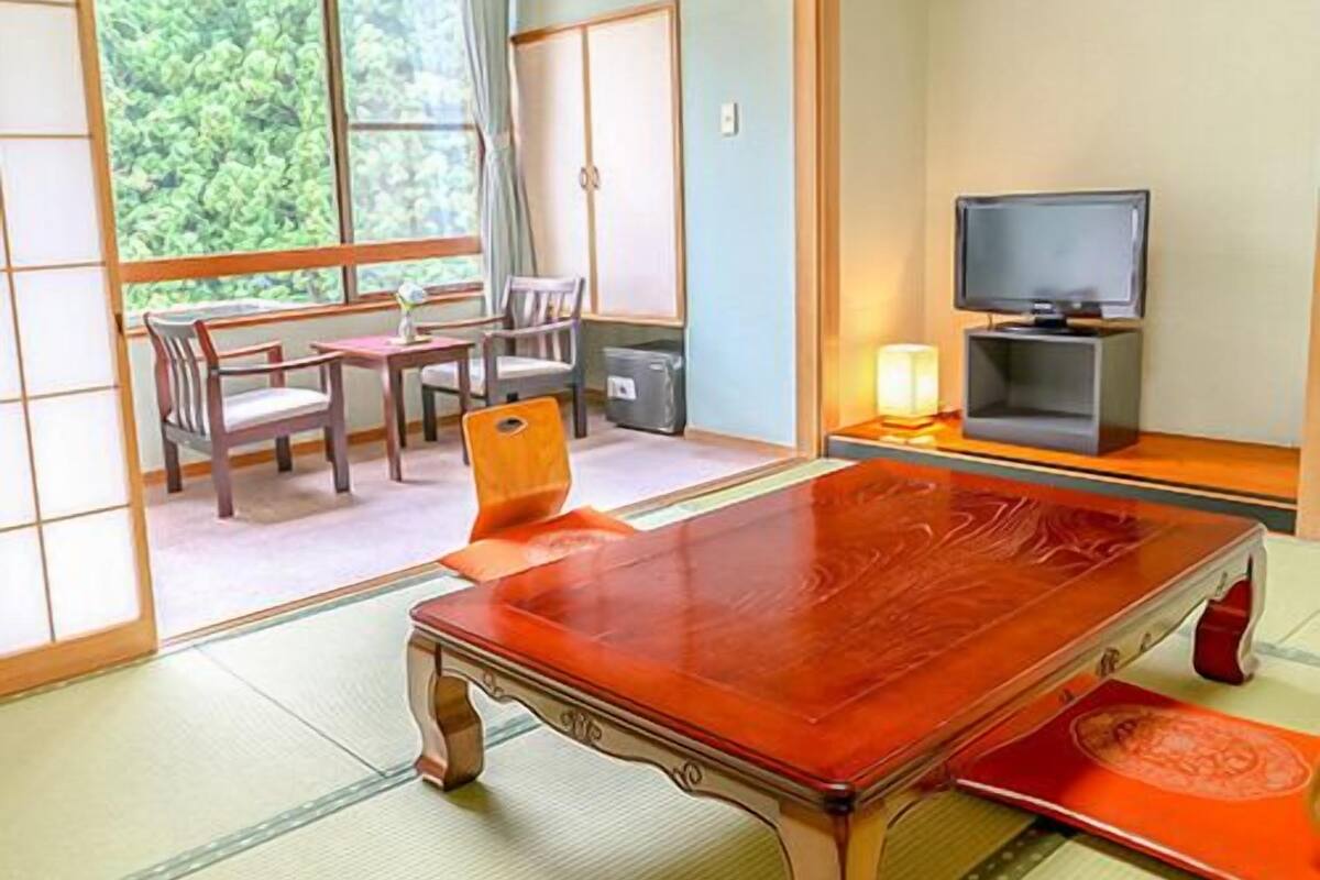 A traditional Japanese room with tatami flooring, a low wooden table, a television, and a view of greenery outside.