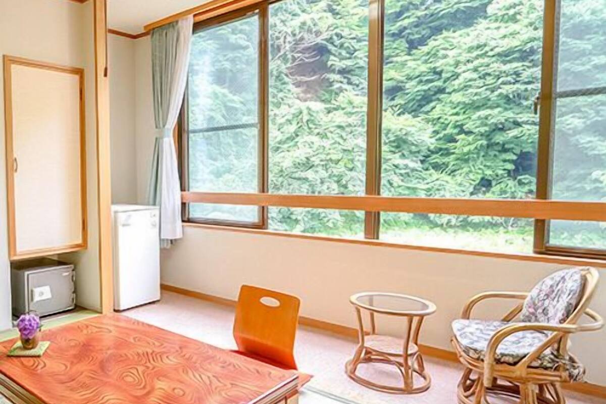 A room with a wooden table, two chairs, a small round table, and a view of greenery through large windows.