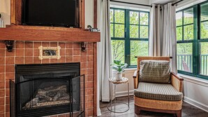 A cozy living room with a fireplace, a comfortable armchair, and a view of greenery through large windows. - Heated Pool Hot Tubs Sauna Gym Deck Mtn Front Appalachian @ Mtn Creek #246/248 (Vernon)