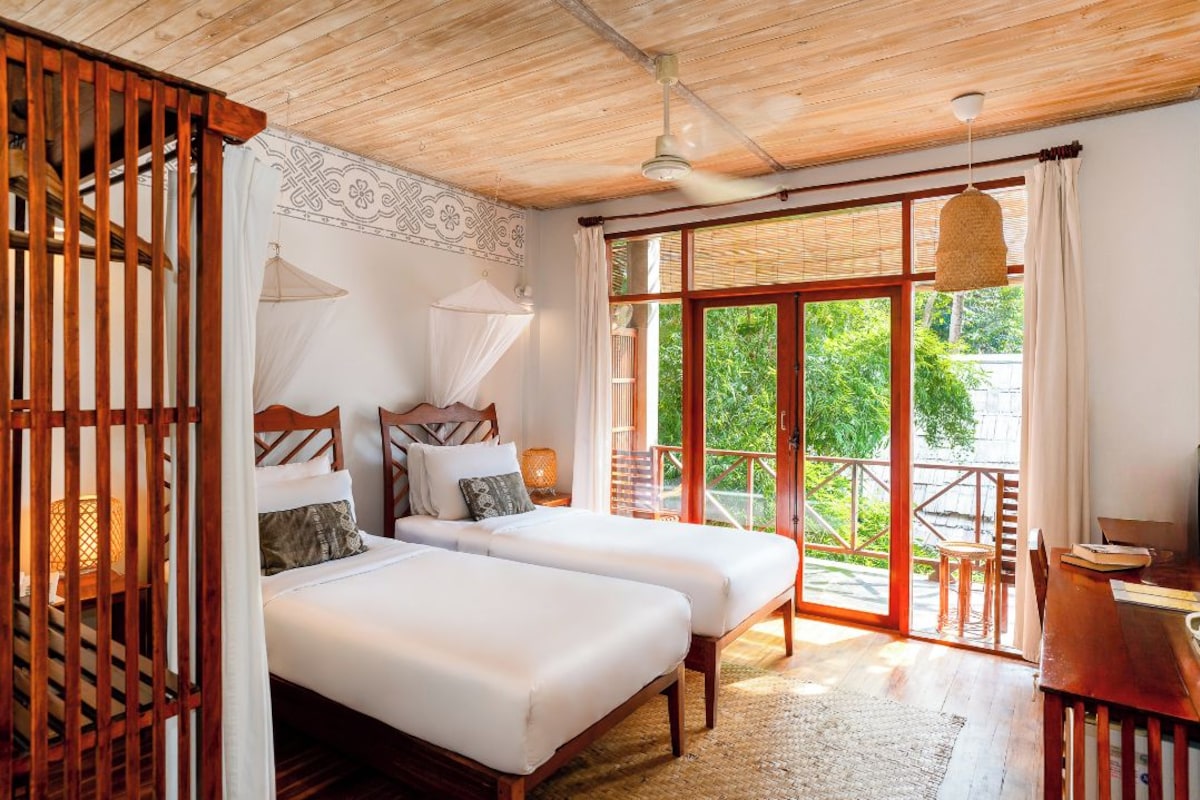 A room with two beds, a wooden ceiling, a balcony with a view of greenery, and a table with chairs.