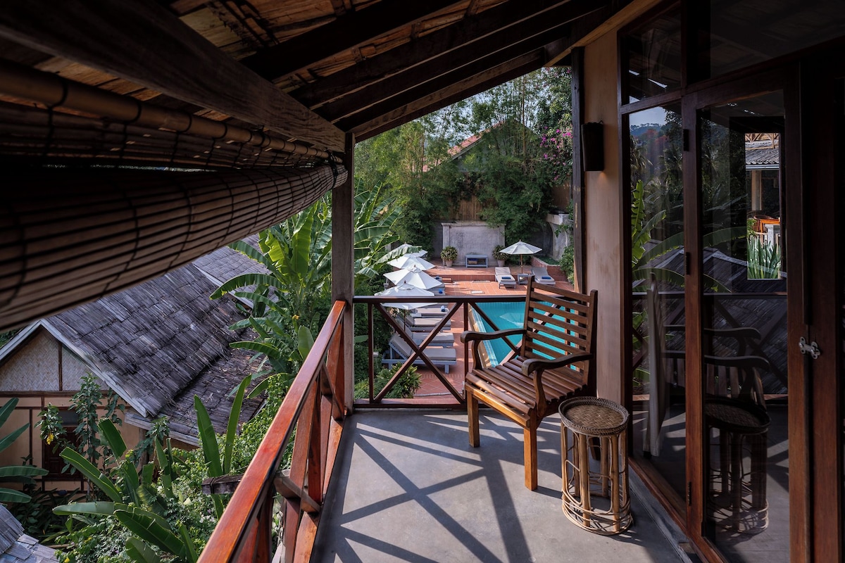 A balcony with a wooden table, chairs, and a small stool. There is a pool in the background, surrounded by greenery.