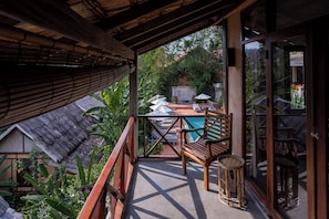 A balcony with a wooden table, chairs, and a small stool. There is a pool in the background, surrounded by greenery. - My Dream Boutique Resort (Luang Prabang)