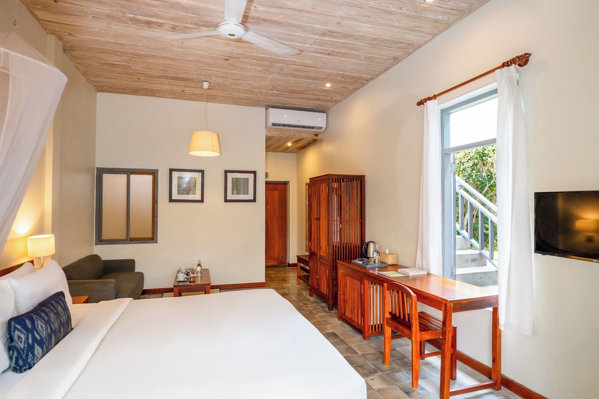 A hotel room with a bed, a desk, a chair, a wardrobe, a ceiling fan, and a window with a view of greenery.