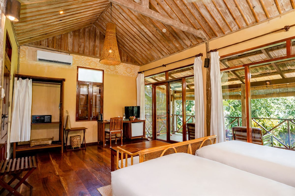 A room with a wooden ceiling, a bed, a desk with a chair, a television, and a large window with curtains.