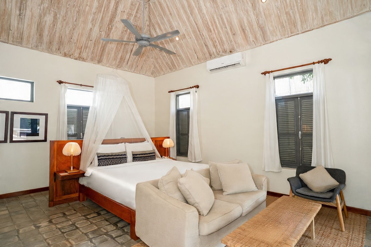 A bedroom with a bed, a sofa, a chair, a ceiling fan, and a window with curtains.