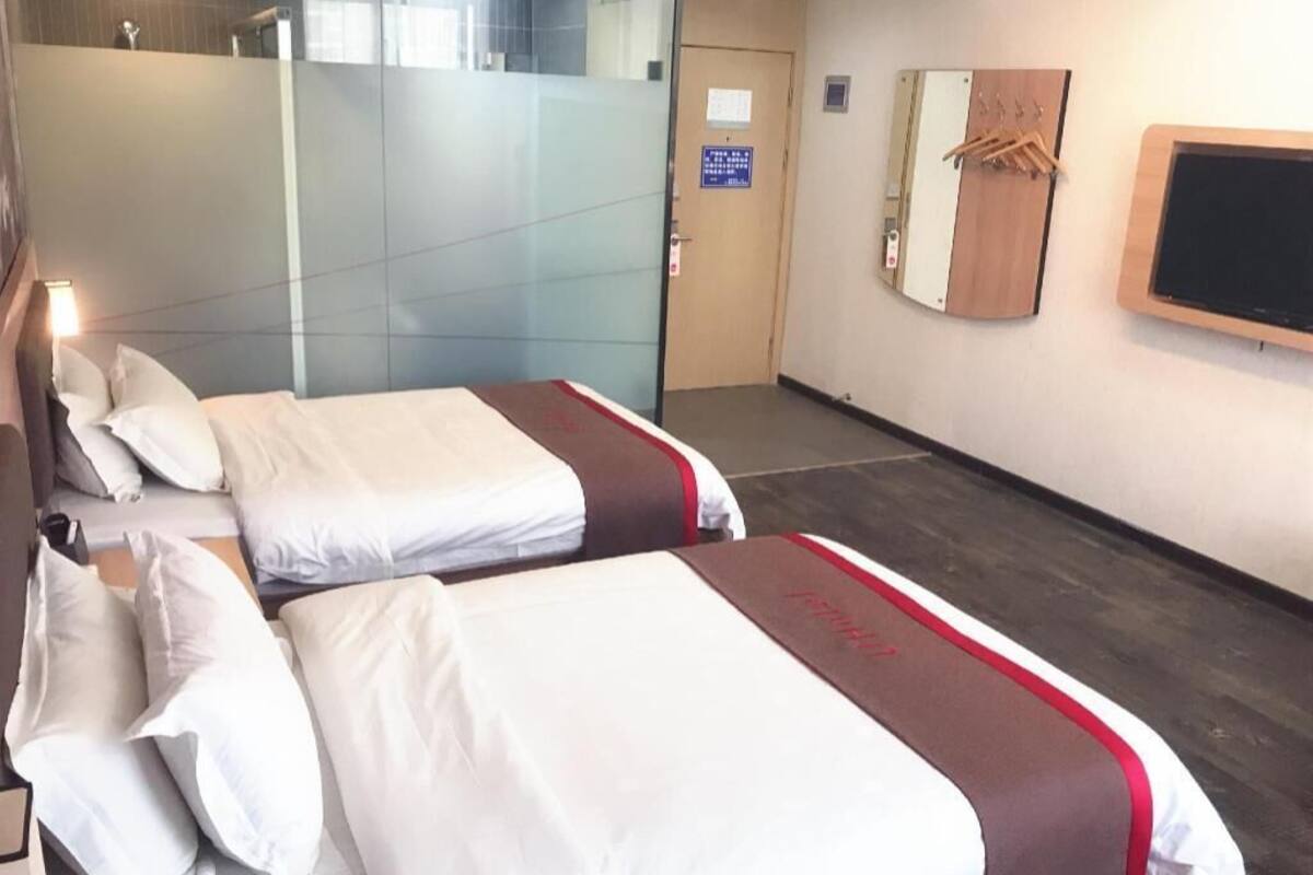 Habitación de hotel con dos camas, zona de ducha, puerta de madera y televisor instalado en la pared.