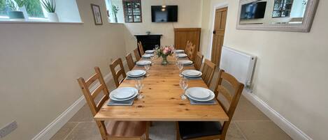 A dining room with a long wooden table set for six, flanked by wooden chairs with black cushions.