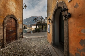 A cobblestone street with a wooden door and a lamp post, leading to a building with a mountain in the background. - Loppia Bay by Wonderful Italy (Bellagio)