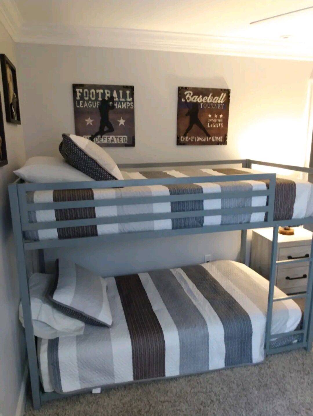 Een stapelbed met streepjesbeddengoed en kussens, een nachtkastje met lades, en twee wanddecoraties met sportthema.