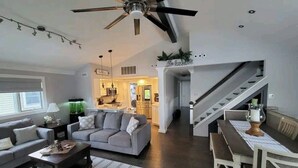 A spacious living room with a gray sofa, a coffee table, and a dining area with a white table and bench. A ceiling fan is visible, and there is a staircase leading to another level. - HOT TUB & Bicycles - Walking distance to the BEACH, bay, restaurants, & shopping (Brigantine)