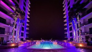 Un área de piscina con una pileta central, rodeada de palmeras e iluminada por luces de colores.