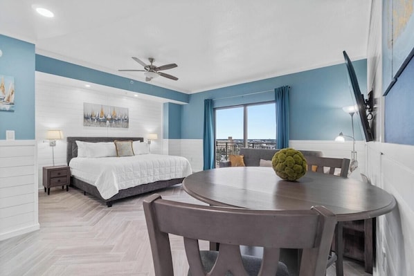 A modern hotel room with a bed, a round dining table, a ceiling fan, and a large window with a city view.
