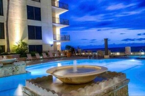 A swimming pool with a central fountain, lounge chairs, and a building with balconies in the background.