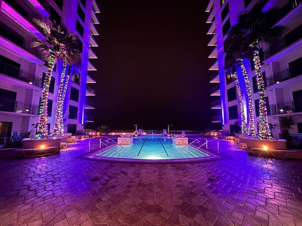 A swimming pool area with a tiled deck, surrounded by palm trees and buildings with balconies.