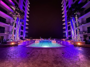 Una piscina con solárium azulejado, rodeada de palmeras y edificios con balcones.