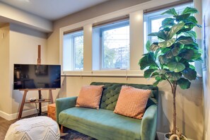 A living room with a green sofa, two cushions, a television on a stand, a large plant, and a window with blinds. - Walkable Portland Studio < 1 Mi to Dtwn! (Portland)