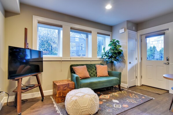 A living room with a large flat-screen TV on a wooden stand, a green sofa with two cushions, a potted plant, and a patterned rug. - Walkable Portland Studio < 1 Mi to Dtwn! (Portland)