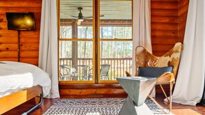 Habitación de cabaña de madera con una cama, una silla con una manta de piel, una mesa y un gran ventanal con vista a muebles de exterior.