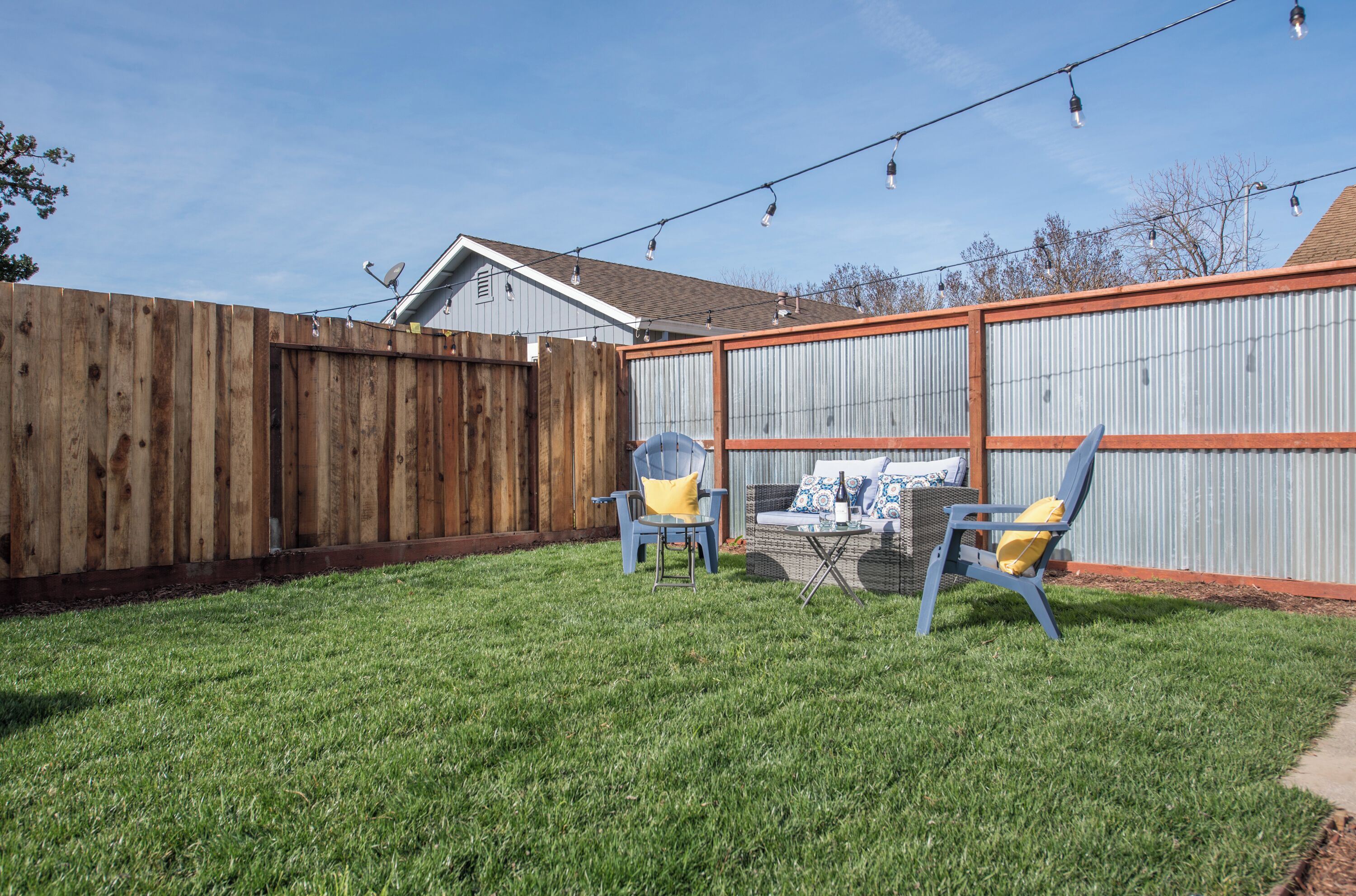 A backyard with a wooden fence, a grassy lawn, and two blue chairs with yellow cushions.