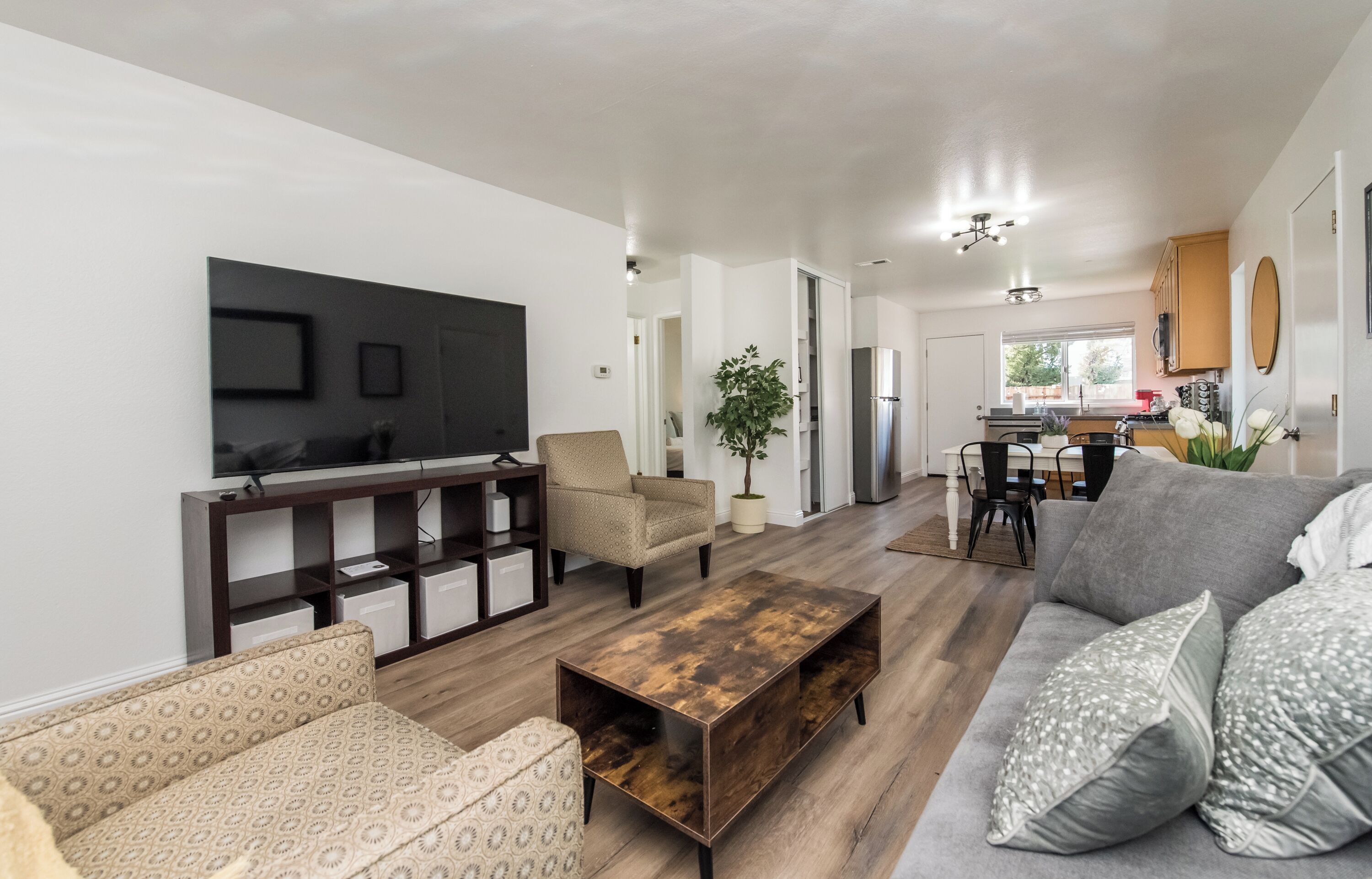 A modern living room with a TV, sofa, coffee table, and a dining area with chairs and a plant.