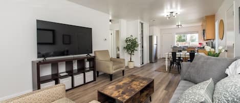 A modern living room with a TV, sofa, coffee table, and a dining area with chairs and a plant.