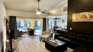 A spacious living room with a large sofa, a coffee table, and a television on a stand. There is a dining area with a table and chairs visible through a glass door. - Heated Pool Home with Southern Exposure! A Tropical Paradise! (Cape Coral)