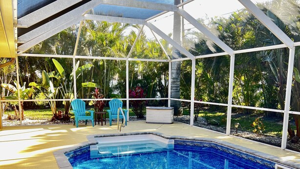 An indoor swimming pool with clear blue water, surrounded by a white structure with glass panels. There are two blue chairs and a small table with a heater. - Heated Pool Home with Southern Exposure! A Tropical Paradise! (Cape Coral)