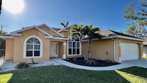 A single-story house with a beige exterior, a garage, and a small front yard with a palm tree and a star-shaped planter. - Heated Pool Home with Southern Exposure! A Tropical Paradise! (Cape Coral)