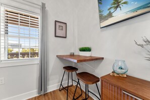 A corner room with a wooden desk, two stools, a framed picture, a small potted plant, and a fishbowl.
